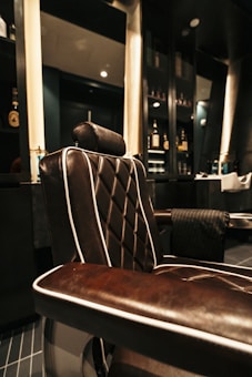 A luxurious barber chair with dark brown leather upholstery featuring white piping, set in an upscale barbershop. The chair is positioned next to shelves containing various bottles, likely grooming products or refreshments. The shop has a modern and elegant interior design, with dark colors and subtle lighting contributing to an overall sophisticated atmosphere.