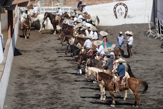 A group of people dressed in traditional attire sit atop horses in a line. They are wearing wide-brimmed hats and colorful clothing, gathered in an arena with a dirt floor. The wall in the background is white with brick elements, and a horseshoe emblem is displayed on it. Some individuals are standing nearby, also wearing similar attire.