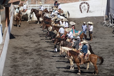 A group of people dressed in traditional attire sit atop horses in a line. They are wearing wide-brimmed hats and colorful clothing, gathered in an arena with a dirt floor. The wall in the background is white with brick elements, and a horseshoe emblem is displayed on it. Some individuals are standing nearby, also wearing similar attire.