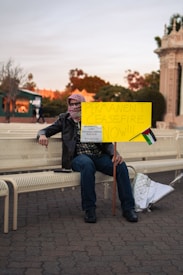 A person wearing a keffiyeh mask and leather jacket is sitting on a bench, holding a yellow protest sign reading 'PERMANENT CEASEFIRE NOW!!!'. A small Palestinian flag is attached to the sign, and there's a piece of silver insulation material on the bench. The background includes some autumn trees and a classical-style structure.