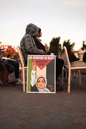 A decorated sign resting against chairs shows an illustration of a person wearing a headscarf, raising a fist holding an olive branch, with text advocating for a ceasefire in Gaza. People are seated behind the sign.