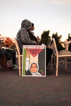 A decorated sign resting against chairs shows an illustration of a person wearing a headscarf, raising a fist holding an olive branch, with text advocating for a ceasefire in Gaza. People are seated behind the sign.