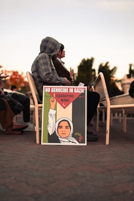 A decorated sign resting against chairs shows an illustration of a person wearing a headscarf, raising a fist holding an olive branch, with text advocating for a ceasefire in Gaza. People are seated behind the sign.