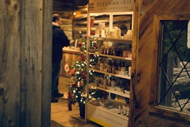A rustic wooden store interior decorated for the holidays with a small Christmas tree adorned with lights and red ornaments beside a shelf displaying skincare products from Beekman 1802. A person stands in the background, browsing items.