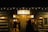 The exterior of a rustic wooden farm shop is warmly lit by outdoor lights. The shop is adorned with a sign that reads 'A Country Christmas Farm Shop'. Several people are seen entering or exiting through the open doors, suggesting a festive, welcoming atmosphere. A 'No Smoking' sign is visible on the right-hand side.