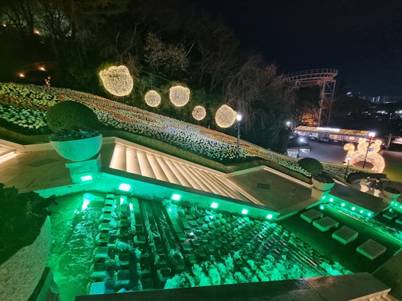 A nighttime scene featuring an illuminated garden with spherical light decorations on a sloping hill. The foreground shows a set of stairs with embedded lighting, leading to a small pool or fountain area lit with green lights. A decorative structure resembling a roller coaster is visible in the background.