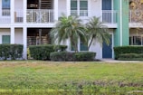 A residential building with a white exterior and a green door. Palm trees and shrubs line the front, while a small lawn is visible in the foreground. The building has an upper balcony and a staircase leading upstairs.