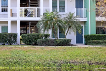 A residential building with a white exterior and a green door. Palm trees and shrubs line the front, while a small lawn is visible in the foreground. The building has an upper balcony and a staircase leading upstairs.