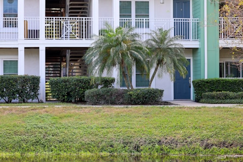 A residential building with a white exterior and a green door. Palm trees and shrubs line the front, while a small lawn is visible in the foreground. The building has an upper balcony and a staircase leading upstairs.