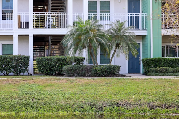 A residential building with a white exterior and a green door. Palm trees and shrubs line the front, while a small lawn is visible in the foreground. The building has an upper balcony and a staircase leading upstairs.