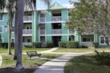 A multi-story apartment building with light green siding and white trim surrounded by well-kept lawns and palm trees. The structure includes open balconies with white railings and a central staircase. A bench and a walking path can be seen in the foreground.
