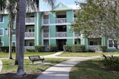 A multi-story apartment building with light green siding and white trim surrounded by well-kept lawns and palm trees. The structure includes open balconies with white railings and a central staircase. A bench and a walking path can be seen in the foreground.