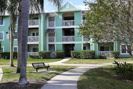 A multi-story apartment building with light green siding and white trim surrounded by well-kept lawns and palm trees. The structure includes open balconies with white railings and a central staircase. A bench and a walking path can be seen in the foreground.