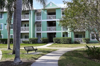 A multi-story apartment building with light green siding and white trim surrounded by well-kept lawns and palm trees. The structure includes open balconies with white railings and a central staircase. A bench and a walking path can be seen in the foreground.
