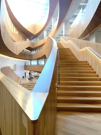 A large, modern interior space featuring a sweeping wooden staircase with clean lines and a smooth finish. The architecture includes curved wooden panels and ample natural light from a skylight. The area seems open and airy, with multiple levels visible.