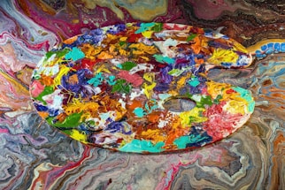 A vibrant artist's palette filled with a variety of acrylic colors is displayed against a swirling, colorful background. The palette itself is covered in thick, textured blobs and strokes of paint in shades of blue, green, orange, red, and yellow, among others. The background features a mesmerizing marbled pattern in a similar color scheme, enhancing the chaotic yet artistic feel.