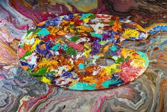 A vibrant artist's palette filled with a variety of acrylic colors is displayed against a swirling, colorful background. The palette itself is covered in thick, textured blobs and strokes of paint in shades of blue, green, orange, red, and yellow, among others. The background features a mesmerizing marbled pattern in a similar color scheme, enhancing the chaotic yet artistic feel.