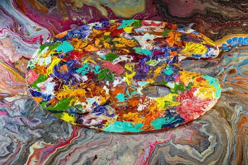 A vibrant artist's palette filled with a variety of acrylic colors is displayed against a swirling, colorful background. The palette itself is covered in thick, textured blobs and strokes of paint in shades of blue, green, orange, red, and yellow, among others. The background features a mesmerizing marbled pattern in a similar color scheme, enhancing the chaotic yet artistic feel.