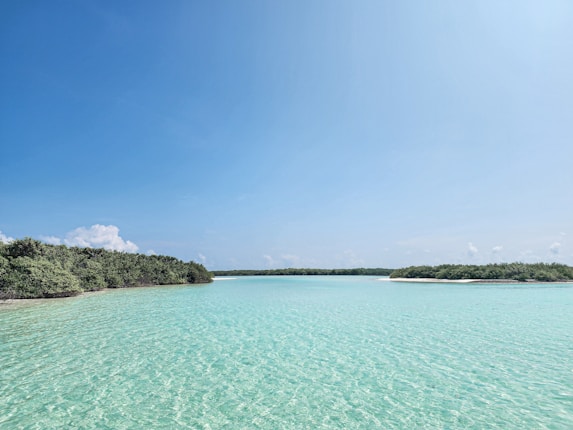 Crystal clear turquoise water stretches across the foreground, merging with a tranquil blue sky. Lush green islands rim the horizon, under a light scattering of white, fluffy clouds.