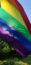 A softball flying over a rainbow-colored field with the LGBT pride flag waving in the background.