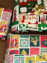 Three wrapped gift boxes featuring festive, multicolored holiday-themed wrapping paper, showcasing patterns of snowmen, Santa Claus, Christmas trees, stockings, and ornaments. A shiny gold bow is attached to one of the presents.