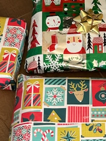 Three wrapped gift boxes featuring festive, multicolored holiday-themed wrapping paper, showcasing patterns of snowmen, Santa Claus, Christmas trees, stockings, and ornaments. A shiny gold bow is attached to one of the presents.