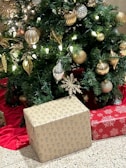 A festive Christmas tree decorated with golden and silver baubles, glittering leaves, and glowing LED lights. Two wrapped gifts sit beneath the tree on a red cloth. One gift is wrapped in brown paper with black tree patterns, and another is wrapped in red paper with white snowflake designs.