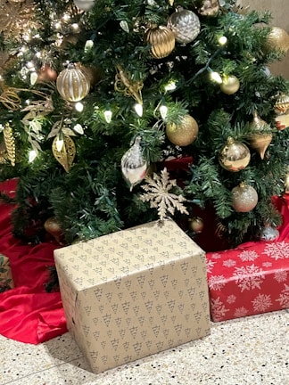 A festive Christmas tree decorated with golden and silver baubles, glittering leaves, and glowing LED lights. Two wrapped gifts sit beneath the tree on a red cloth. One gift is wrapped in brown paper with black tree patterns, and another is wrapped in red paper with white snowflake designs.
