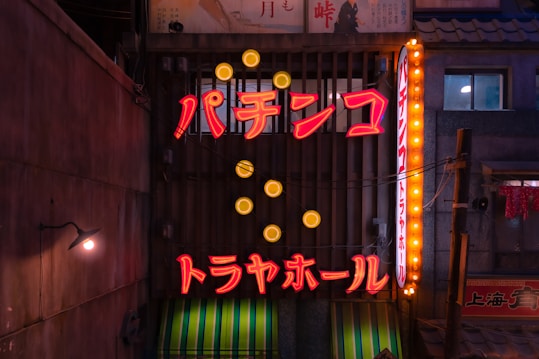 A neon-lit sign in Japanese script displayed on a building, surrounded by circular yellow lights. The setting appears to be a narrow urban alley with muted, shadowy surroundings and additional signage in the background.