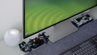 A computer monitor with a sleek design sits on a desk, displaying a green gradient wallpaper. In front of the monitor, there are two miniature Formula 1 race car models. To the left, a spherical smart speaker with a mesh covering is placed on the desk, next to a keyboard.