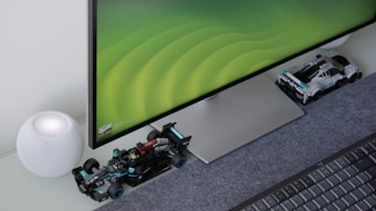 A computer monitor with a sleek design sits on a desk, displaying a green gradient wallpaper. In front of the monitor, there are two miniature Formula 1 race car models. To the left, a spherical smart speaker with a mesh covering is placed on the desk, next to a keyboard.