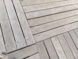 Weathered wooden planks with an interlocking pattern and visible screws, showing signs of aging and wear, possibly part of a boardwalk or deck.