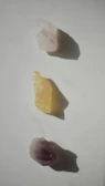 Three raw crystals placed in a vertical line on a light surface. The top crystal is translucent with a white hue, the middle one is opaque with a yellow tint, and the bottom crystal displays a purple shade. Each crystal casts a distinct shadow on the surface.