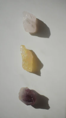 Three raw crystals placed in a vertical line on a light surface. The top crystal is translucent with a white hue, the middle one is opaque with a yellow tint, and the bottom crystal displays a purple shade. Each crystal casts a distinct shadow on the surface.