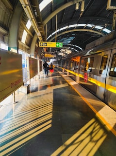 A modern metro station platform with an approaching train on the track. The platform is brightly lit with natural sunlight filtering through the arched roof. Shadow patterns from the roof structure create dynamic lines on the platform. Two people are walking towards the exit, accompanied by prominent sign boards indicating directions.