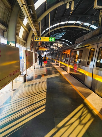 A modern metro station platform with an approaching train on the track. The platform is brightly lit with natural sunlight filtering through the arched roof. Shadow patterns from the roof structure create dynamic lines on the platform. Two people are walking towards the exit, accompanied by prominent sign boards indicating directions.
