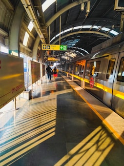 A modern metro station platform with an approaching train on the track. The platform is brightly lit with natural sunlight filtering through the arched roof. Shadow patterns from the roof structure create dynamic lines on the platform. Two people are walking towards the exit, accompanied by prominent sign boards indicating directions.