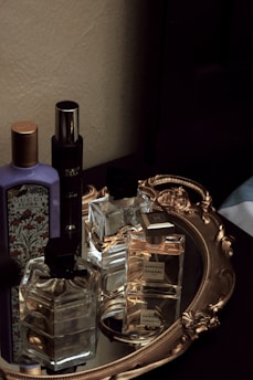 A collection of perfume bottles is arranged on an ornate, oval gold-toned tray with a mirrored base. The tray is placed on a dark surface against a light-colored wall. Several glass bottles of various shapes and sizes are visible, with some featuring recognizable brand labels.
