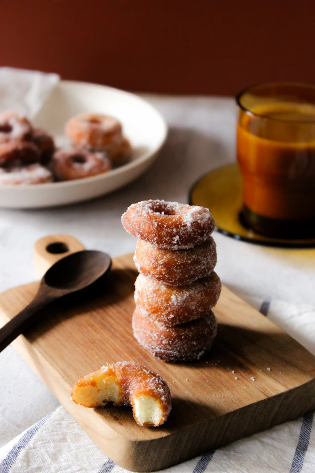 A stack of small sugar-dusted donuts on a wooden board with a spoon beside it