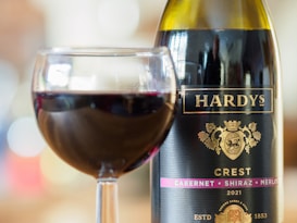 A large bottle of Hardy's Crest wine labeled with a crest insignia and 2021 Cabernet Shiraz Merlot blend stands next to a filled glass of red wine. The background is softly blurred, creating a bokeh effect with various light colors.