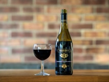A bottle of red wine with the label 'Hardy's' stands next to a filled wine glass on a wooden surface. The background features a blurred brick wall, adding a rustic atmosphere.