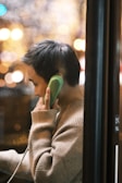 A person with short hair is using a bright green public telephone. They are dressed in a light beige sweater and are seen from the side through a window with bokeh lighting effects in the background, suggesting a nighttime or urban environment.