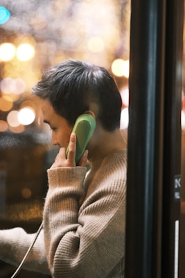 A person with short hair is using a bright green public telephone. They are dressed in a light beige sweater and are seen from the side through a window with bokeh lighting effects in the background, suggesting a nighttime or urban environment.
