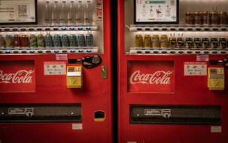 Two adjacent red vending machines stocked with a variety of beverages, including bottled water, soft drinks, and canned coffee. The machines prominently feature the Coca-Cola logo. Both machines have payment options displayed, including cash and QR code payments.