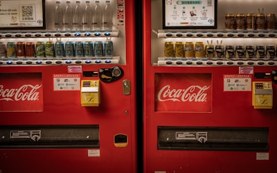Two adjacent red vending machines stocked with a variety of beverages, including bottled water, soft drinks, and canned coffee. The machines prominently feature the Coca-Cola logo. Both machines have payment options displayed, including cash and QR code payments.
