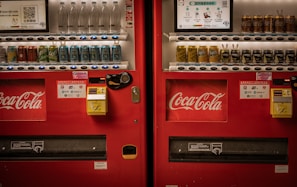 Two adjacent red vending machines stocked with a variety of beverages, including bottled water, soft drinks, and canned coffee. The machines prominently feature the Coca-Cola logo. Both machines have payment options displayed, including cash and QR code payments.