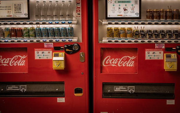 Two adjacent red vending machines stocked with a variety of beverages, including bottled water, soft drinks, and canned coffee. The machines prominently feature the Coca-Cola logo. Both machines have payment options displayed, including cash and QR code payments.