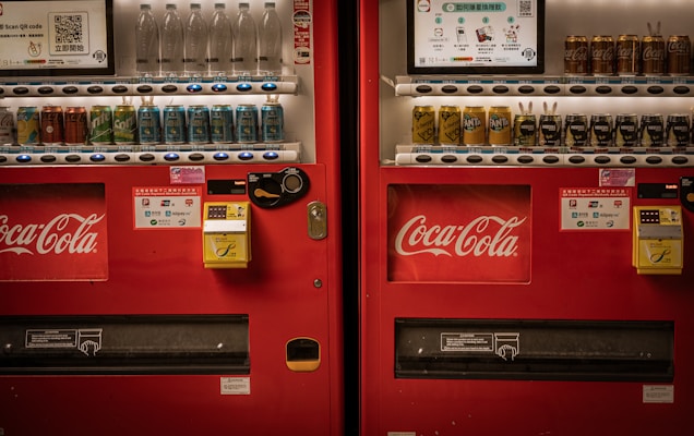 Two adjacent red vending machines stocked with a variety of beverages, including bottled water, soft drinks, and canned coffee. The machines prominently feature the Coca-Cola logo. Both machines have payment options displayed, including cash and QR code payments.