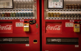 Two adjacent red vending machines stocked with a variety of beverages, including bottled water, soft drinks, and canned coffee. The machines prominently feature the Coca-Cola logo. Both machines have payment options displayed, including cash and QR code payments.