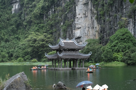 A traditional pagoda-style structure stands in a serene body of water surrounded by lush greenery and towering rock cliffs. People wearing conical hats are paddling boats on the calm water, while some hold umbrellas.